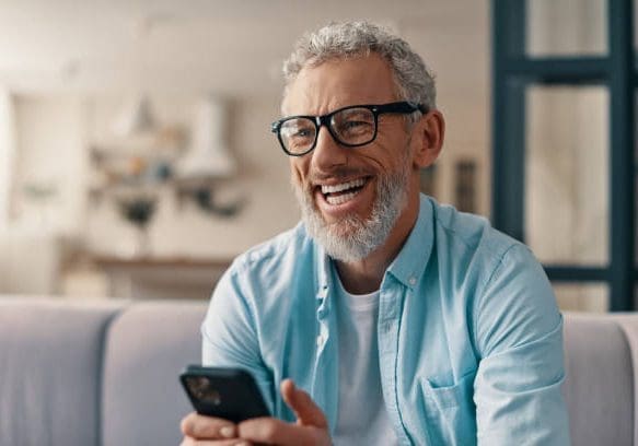 Cheerful senior man in casual clothing and eyeglasses using smart phone while sitting on the sofa at home
